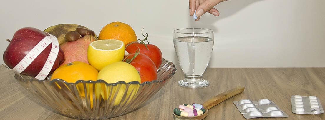suplementos dietéticos: mano echando un suplemento dietético a una copa con agua y diversas frutas dentro de un recipiente de cristal
