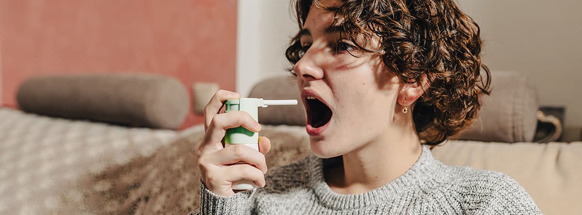 Adolescente, , con un cálido suéter de lana, sentada en un sofá en casa, con dolor de garganta, salpica medicina con spray para la garganta. 